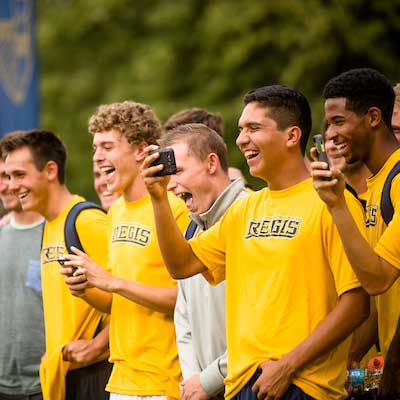 group of male students in gold Regis t-shirts laugh and take pictures outdoors