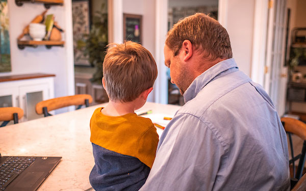 man sits at a counter strewn with books, papers and laptop while holding small child