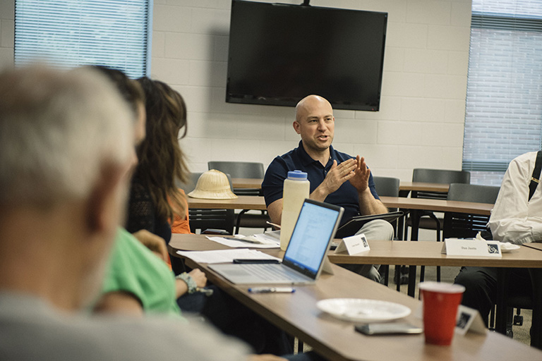 students sit around a table in a classroom with laptops and notebooks, listening to man speak