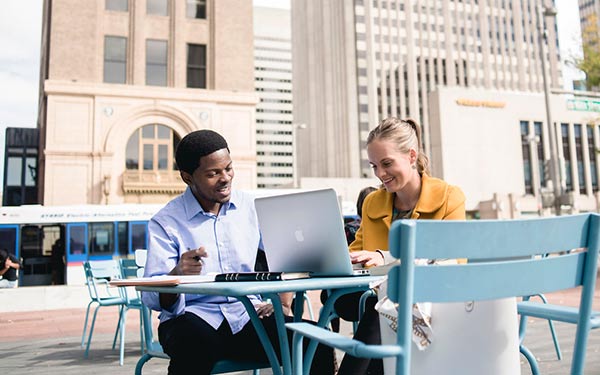 A man and woman sit at an outdoor table, writing on a notebook and laptop