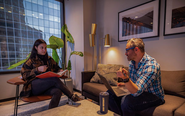 young professional woman reads a book and converses with young professional man who is working on a laptop