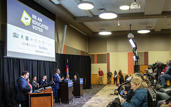 Mayoral candidates sit or stand at the front of a large room full of debate attendees