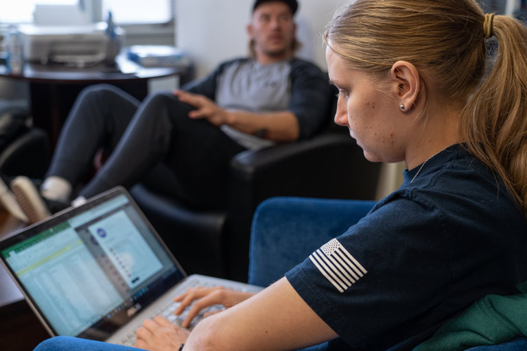 Student in an Air Force tee using a laptop to review course work in a campus lounge with another student in the background.