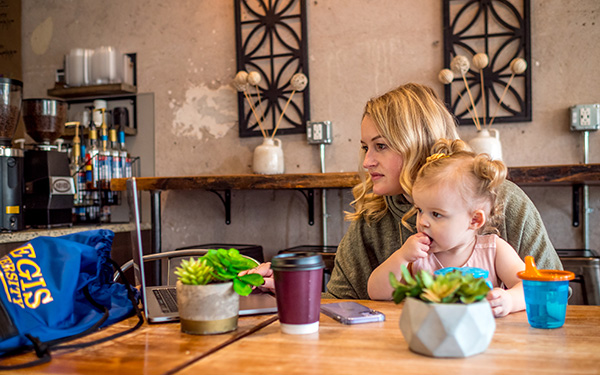 young woman works on laptop while tending her baby and sitting in a coffee shop