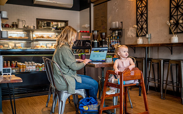 a woman working on a laptop sits beside a toddler in a highchair at a coffee shop table