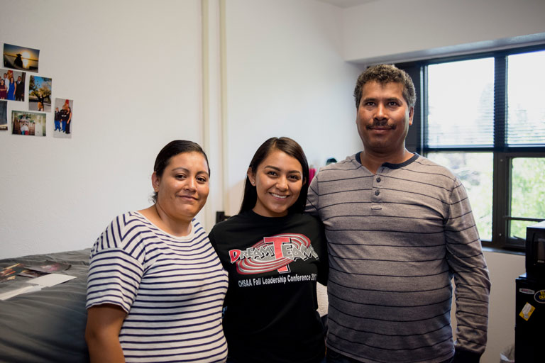 Student stands in the middle of both parents while standing in a dorm that is yet to be completely moved into