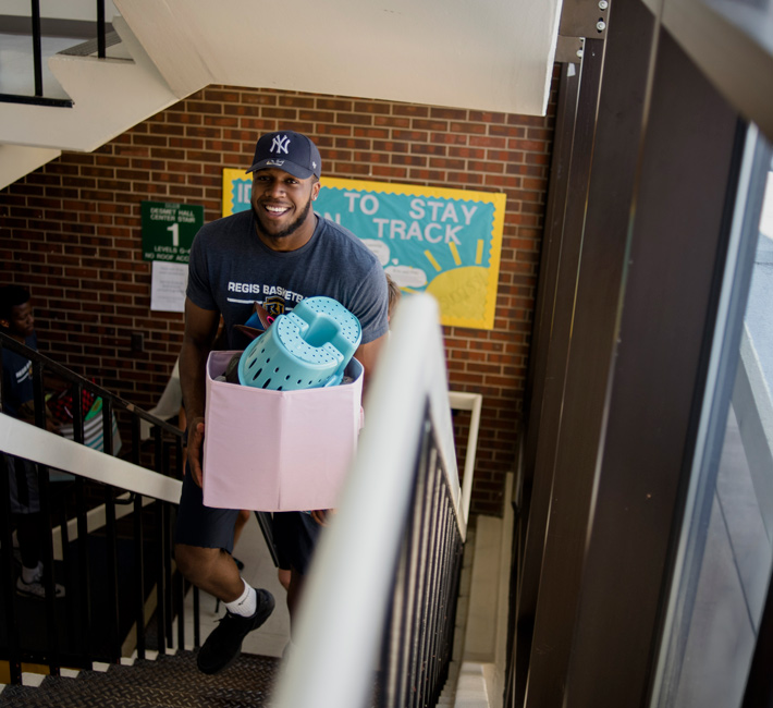 Student walking up the stairs in Desmet Hall carrying a basket during move-in day, smiling with enthusiasm