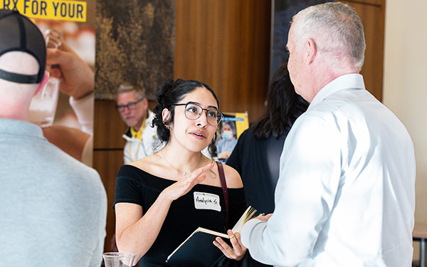 a young woman wearing a name tag reads from a notebook and talks to an older man at a networking event on the Regis University campus