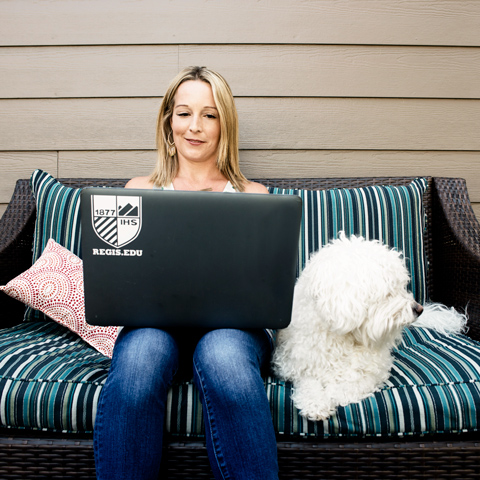 An adult student studying from home sitting on an outdoor couch with her fluffy white dog
