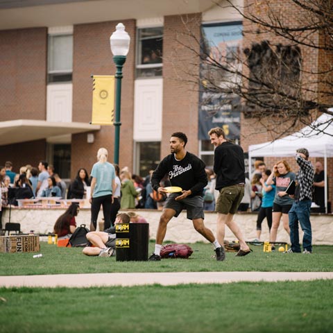 Student throwing a frisbee across the Regis Common with many students mingling in the background.