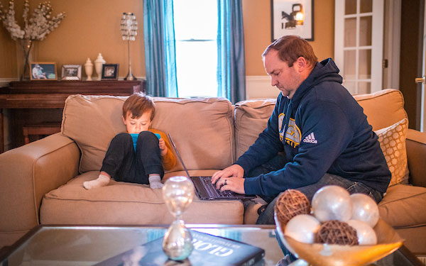man works on a laptop sitting on a sofa beside a small child looking at a tablet