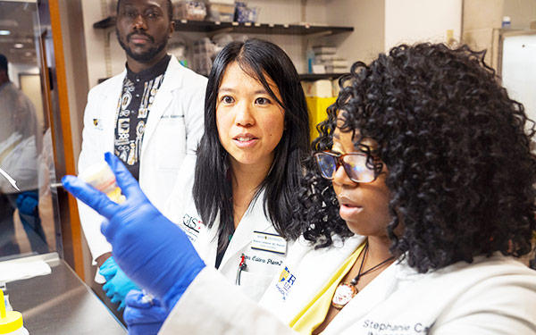 a pharmacy instructor and two students, all in white coats, stand in a pharmacy classroom and watch intently as one student fills a syringe from a bottle with her gloved hand