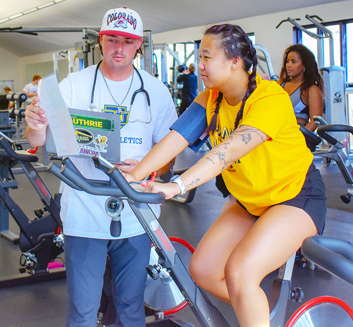 a personal trainer consults with a client who is riding a stationary bike  in the RegisCARES clinic on the Northwest Denver campus