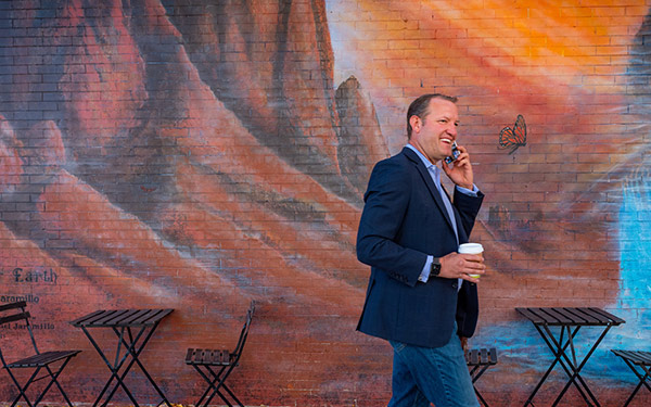 a man wearing a blazer and jeans talks on a phone while standing in front of a colorful mural with a monarch butterfly