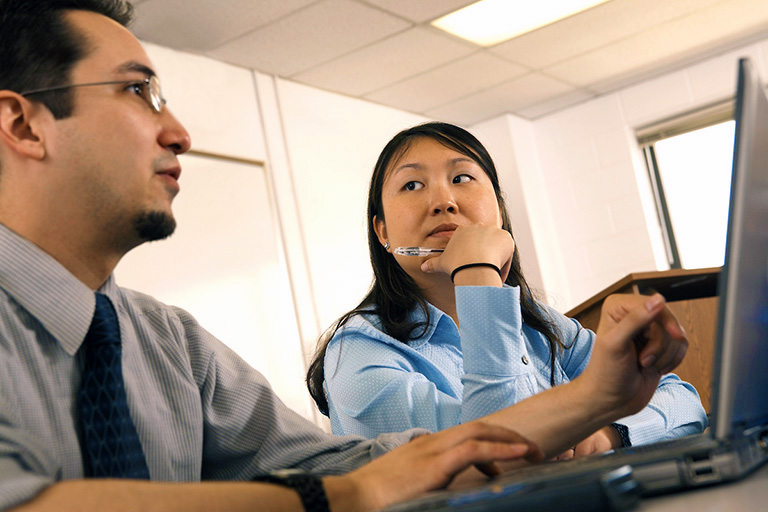 a man and woman in professional attire sit at a desk talking and working on a laptop in a classroom