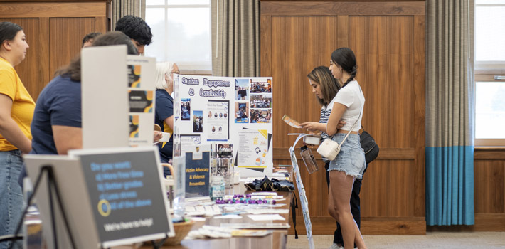 A student and their parents meet with regis staff indoors while looking at poster boards set up explaining the department