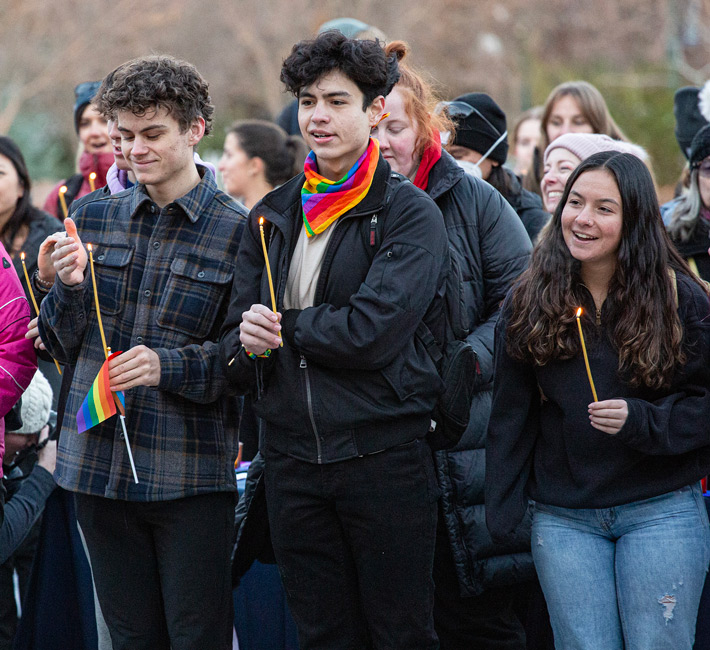Students gather at a queer solidarity march, holding lit candles and small rainbow flags during an outdoor event on campus.