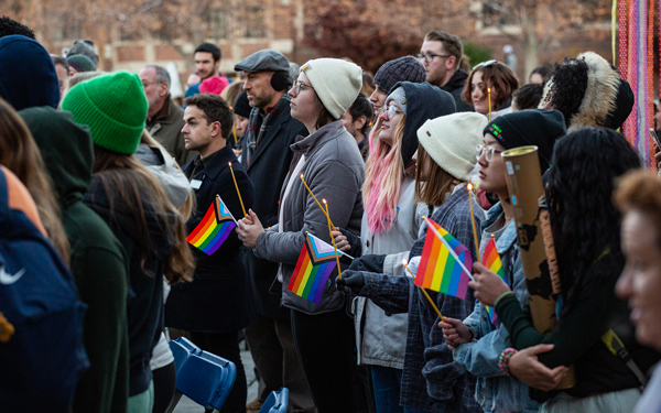 Group of people gathered at an outdoor queer solidarity march, holding candles and Progress Pride flags while dressed in warm clothing