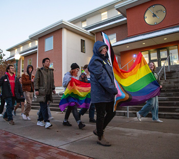 Students participate in a queer solidarity march on campus at dusk, holding rainbow and progress pride flags while walking past a brick building.