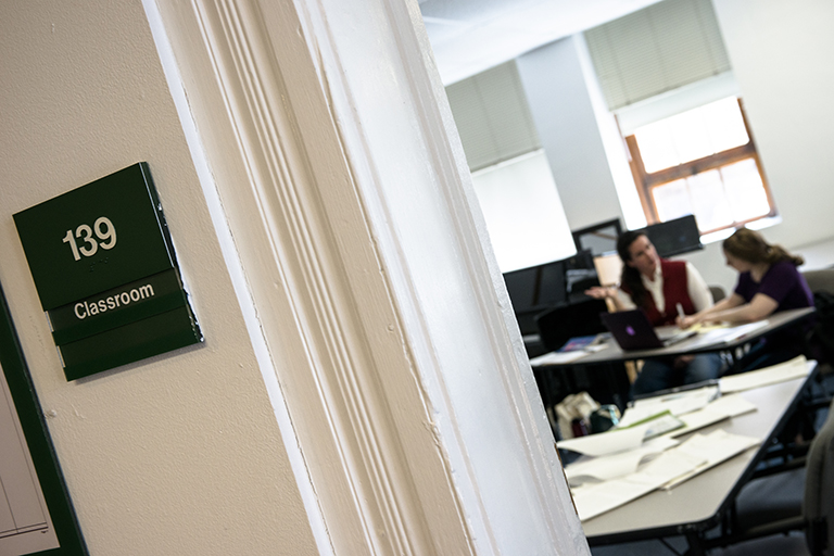 sign reading Classroom 139 on the wall outside a room where a teacher and student sit conversing at desks