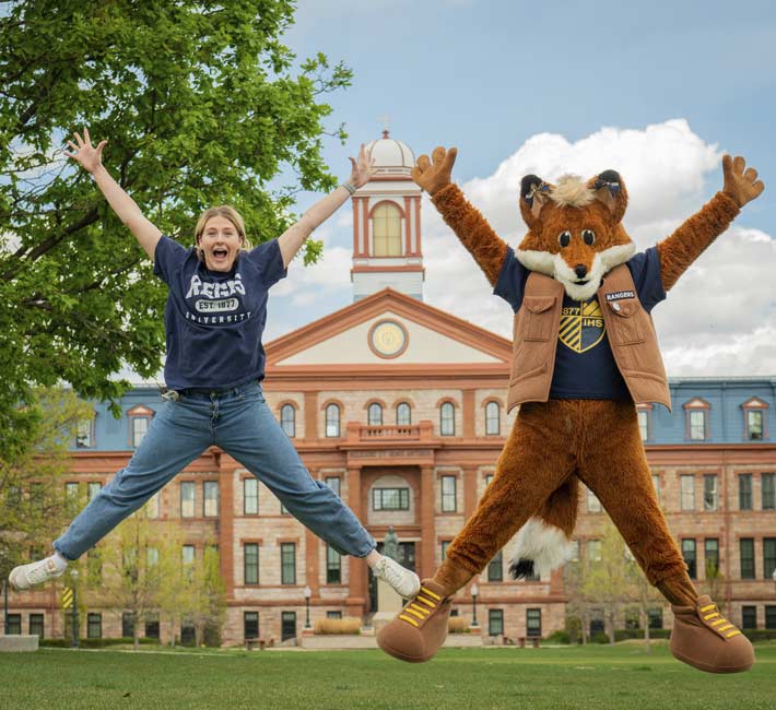 A student on the left and Regi on the right both jump in the air with their hands out and legs out. Main hall and its cupola can be seen right behind them