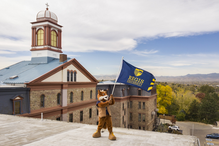 Regi the Ranger mascot waving a Regis flag atop a building with main hall and the Rockies in the background