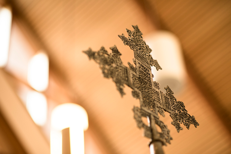 Ornate metal cross with intricate patterns, set against a blurred wooden ceiling background