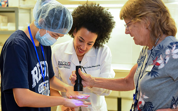 a camper, faculty member and staff member examine the camper's hands with a blue light flashlight to see how much bacteria remains after using hand sanitizer at the Pharmacy Summer Camp on the Northwest Denver campus.