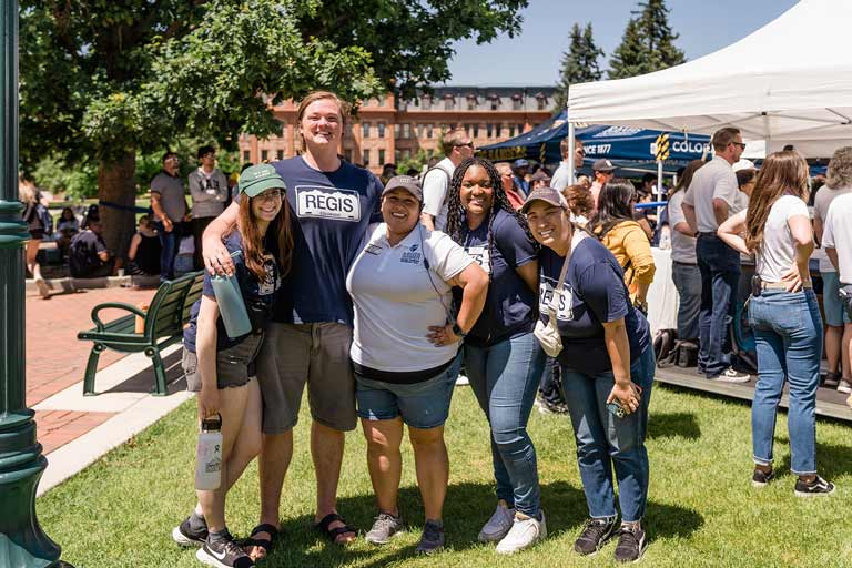 Five individuals stand side by side smiling at the camera during an outdoor admissions event on the Regis quad