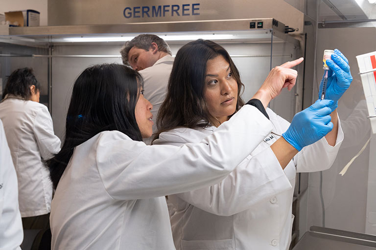 a faculty member in a white coat instructs a pharmacy student as she practices measuring an injection in the pharmacy lab on the Regis campus