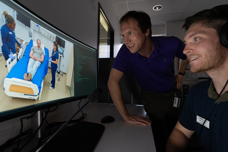 A Regis healthcare student and faculty member watch on a screen as classmates interact with a patient at the Interprofessional Health Site in Thornton, Colorado