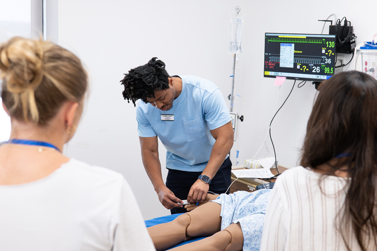 In the clinical simulation lab, a Regis healthcare student works with a patient while faculty and classmates observe