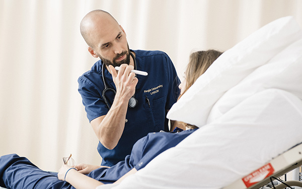 nursing student wearing scrubs uses a flashlight to examine a patient's eyes in the Regis nursing sim lab