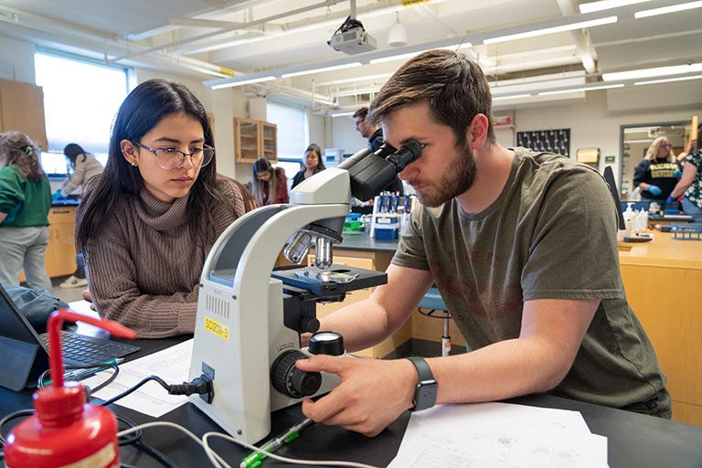 Biology student looks through a microscope while his lab partner takes notes