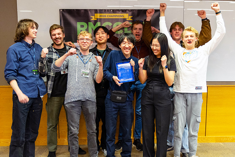 A team of 9 students from CU Boulder hold up the winners plaque while standing in front of a banner that says 2026 Champions RMCCDC