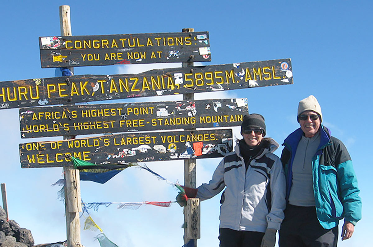 Two people stand in front of a sign on a mountain