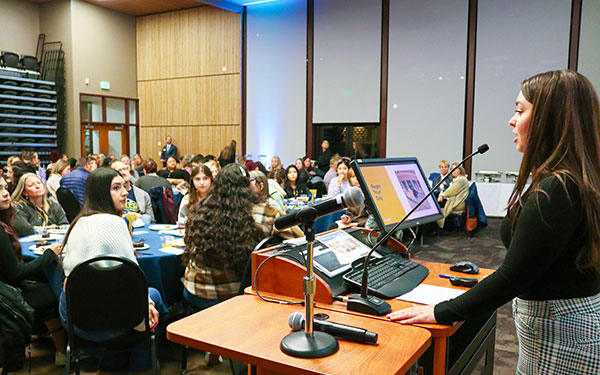 an admissions counselor stands behind a podium delivering a presentation in front of a large room with tables where prospective students and their guests are seated listening