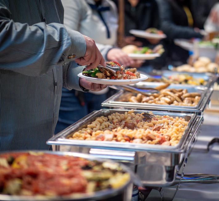 Person stands in front of a table of food and is picking up more food for their plate with tongs