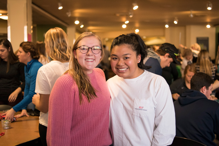 Two students smiling together while others gather and work in the background at a campus event