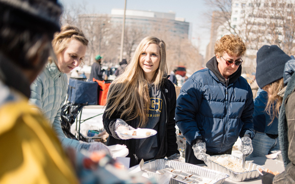 Regis University students volunteer at an outdoor food distribution event, serving meals to people on a cold day in an urban setting.
