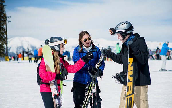 three Regis students holding skis and a snowboard and wearing winter sports gear stand together on the slopes of a snowy ski resort with a crowd of people behind them and the snowy mountains in the background