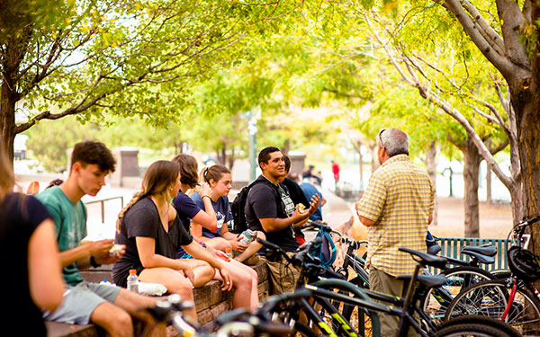 students sit on a wall outdoors in Denver near their parked bikes as some chat with a faculty member during an Outdoor Excursion during Orientation weekend