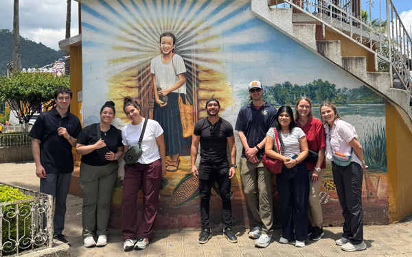 A group of Regis students stand smiling in front of a colorful mural depicting a radiant figure with rays extending outward