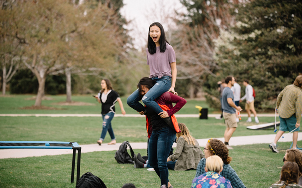 Student sitting on another student’s shoulders on the campus common lawn, smiling and engaging with peers