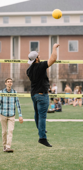 Students playing a volleyball on the commons with one student jumping in the air, mid-spike