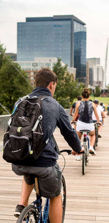 backshot of student riding a bike across a pedestrian bridge in downtown Denver, with skyscraper buildings and trees in the distance