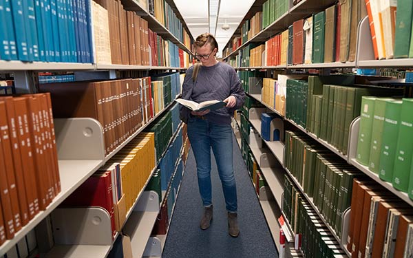 woman stands among the library stacks reading a book