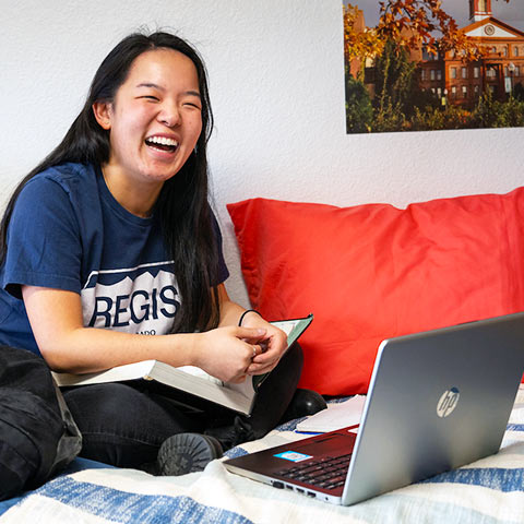 a happy student studies with books and a laptop while sitting on her bed in her dorm room