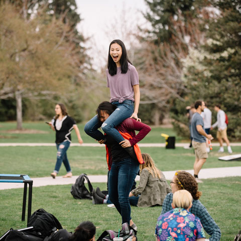 One student sitting on another’s shoulders while others gather on a Regis campus lawn.