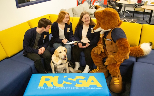 Student leaders meeting with the campus mascot, joined by a service dog, in a campus lounge.